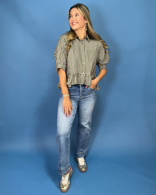 A woman modeling a black and white gingham checkered blouse with puff sleeves and a ruffled hem, paired with blue jeans and metallic wedge sneakers