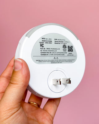Hand holding a white electronic device with visible branding and certification labels against a pink background