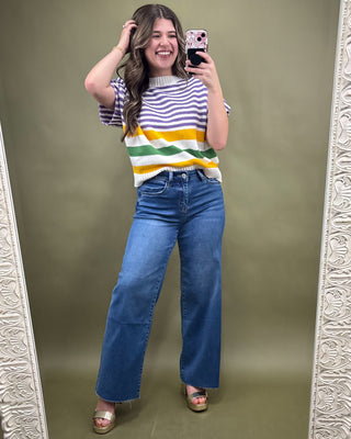 Woman taking a mirror selfie wearing a colorful striped shirt and blue jeans on a green background.