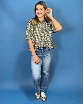 A woman modeling a black and white gingham short-sleeve blouse with a ruffled hem, paired with light-wash blue jeans and silver sneakers