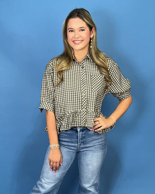 A woman modeling a short-sleeve button-up shirt with a brown and white gingham check pattern and a ruffled hem, paired with blue jeans