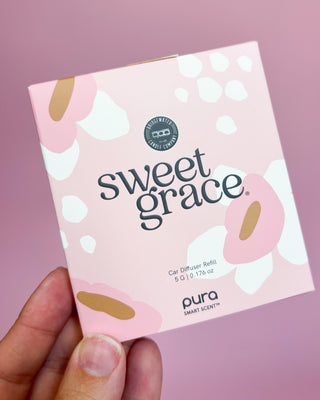 Pura Sweet Grace car diffuser refill packaging held by a hand against a pink background