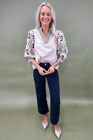 Woman wearing a white blouse with floral patterns, navy blue jeans, and white shoes on a plain background