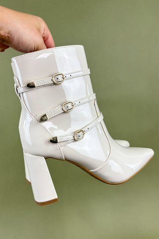 White patent leather boot with buckles held by a hand against a green background