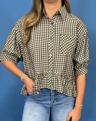 A woman wearing a brown and beige gingham check button-down shirt with a ruffled hem and three-quarter sleeves, paired with blue jeans