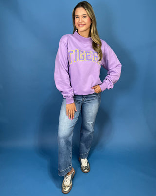 A woman modeling a lavender purple sweatshirt with 'TIGER' printed in gold across the chest, paired with blue jeans and metallic sneakers