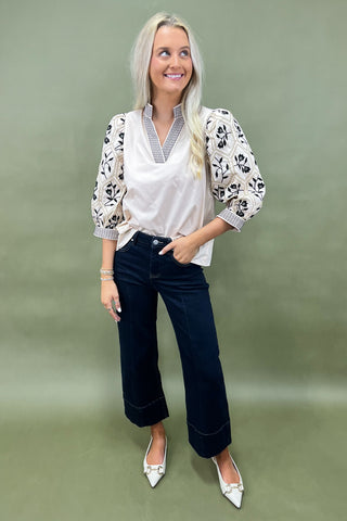 Woman wearing a white blouse with floral patterns, navy blue jeans, and white shoes on a plain background