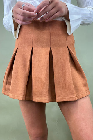 Person wearing a brown pleated skirt with a white top on a light green background