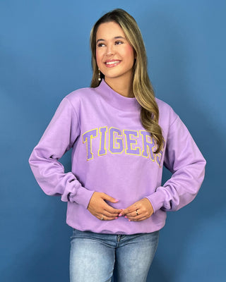 A woman modeling a lavender purple crewneck sweatshirt with 'TIGERS' printed across the chest in yellow and blue varsity lettering