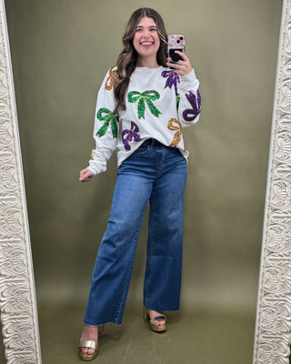 Woman taking a mirror selfie wearing a white sweater with colorful designs and blue jeans.