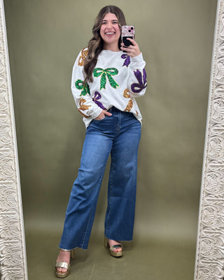 Woman taking a mirror selfie wearing a white sweater with colorful designs and blue jeans.