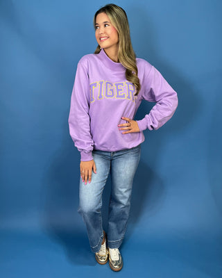 A woman modeling a lavender purple sweatshirt with 'TIGER' printed in yellow varsity letters across the chest, paired with blue jeans and white sneakers
