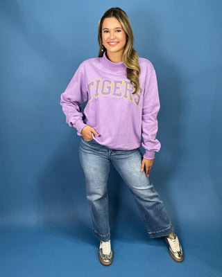 A woman modeling a lavender purple crewneck sweatshirt with 'TIGERS' printed in gold across the chest, paired with blue jean