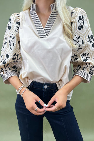 Woman wearing a white blouse with floral patterns, navy blue jeans, and white shoes on a plain background