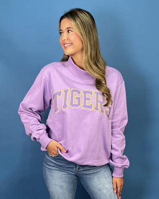 A woman wearing a lavender purple sweatshirt with 'TIGERS' printed across the chest in yellow and blue outline lettering, paired with blue jeans