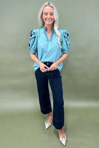 Woman wearing a blue embroidered blouse and dark jeans on a green background