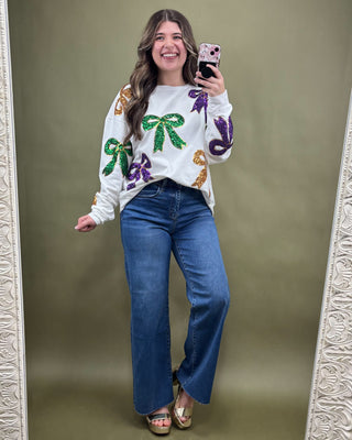 Woman taking a mirror selfie wearing a colorful sweater with bow designs and blue jeans on a green background.