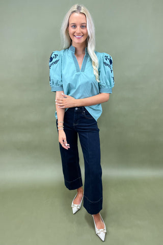 Woman wearing a blue embroidered blouse and dark jeans on a green background