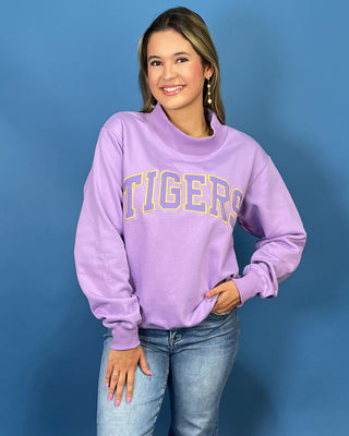 A woman wearing a lavender purple crewneck sweatshirt with 'TIGERS' printed in yellow and white arched letters across the chest