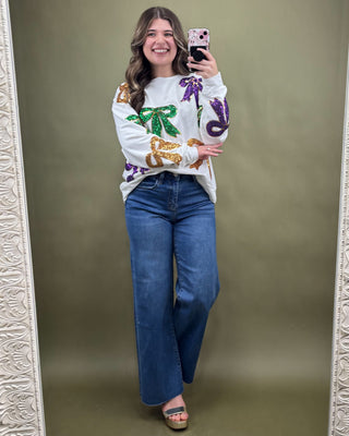 Woman taking a mirror selfie wearing a white sweater with colorful designs and blue jeans on a plain background.