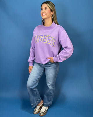 A woman modeling a lavender purple crewneck sweatshirt with 'TIGERS' printed across the chest in yellow and blue varsity lettering, paired with blue jeans and metallic shoes