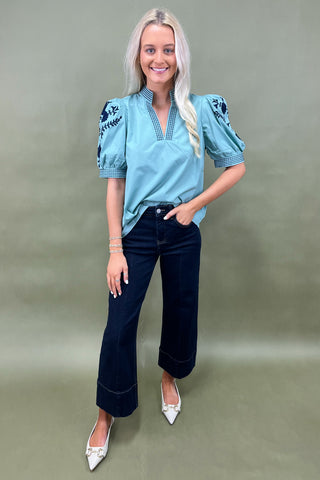 Woman wearing a blue embroidered blouse and dark jeans on a green background