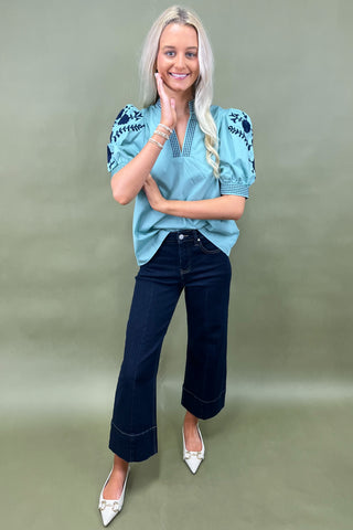 Woman wearing a blue embroidered blouse and dark jeans on a green background
