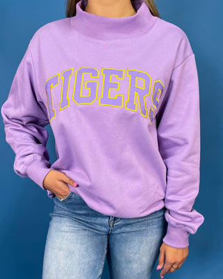A person wearing a lavender purple sweatshirt with 'TIGERS' printed across the chest in yellow and blue outline lettering