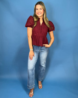 A woman modeling a burgundy eyelet lace short-sleeve blouse with puff sleeves and a ruffled hem, paired with light-wash blue jeans and beige sandal