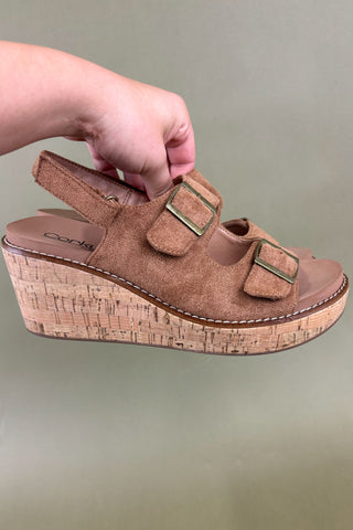 Brown wedge sandal with cork sole held by a hand against a neutral background