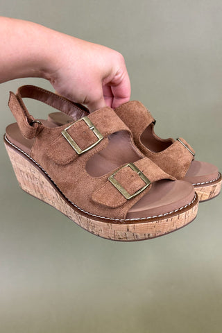 Brown wedge sandals with cork sole held by a hand against a green background