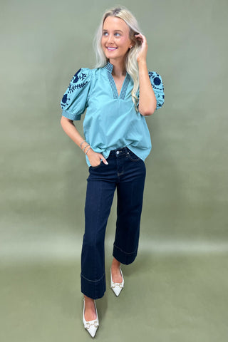 Woman wearing a blue embroidered blouse and dark jeans on a green background