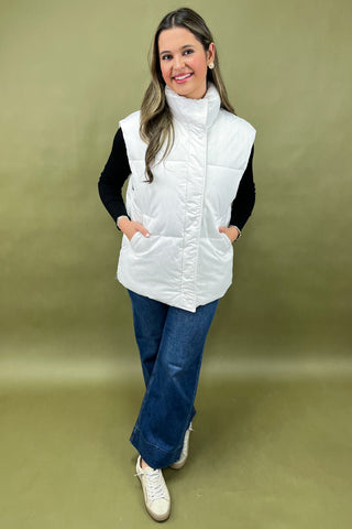 Woman wearing a white puffer vest over a black long-sleeve shirt with blue jeans against a beige background