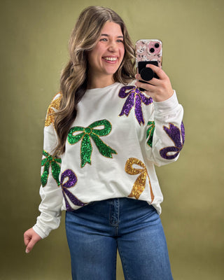 Woman wearing a white sweater with colorful sequin designs, taking a selfie against a green background.