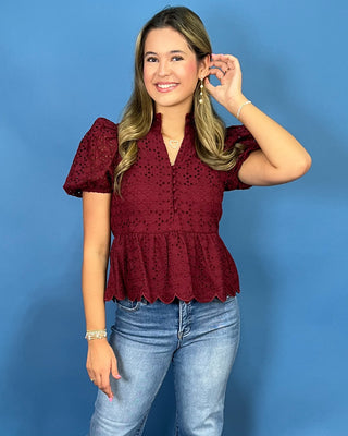 A woman modeling a burgundy eyelet lace short-sleeve blouse with puff sleeves and a ruffled hem, paired with light-wash blue jeans and beige sandal