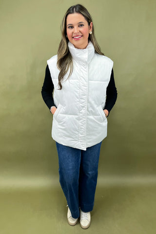 Woman wearing a white puffer vest over a black long-sleeve shirt on a green background