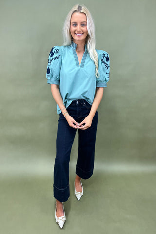 Woman wearing a blue embroidered blouse and dark jeans on a green background