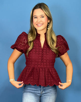 A woman modeling a burgundy eyelet lace peplum top with puff sleeves, paired with blue jeans, against a solid blue studio background.