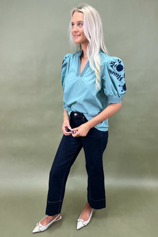 Woman wearing a blue embroidered blouse and dark jeans on a green background