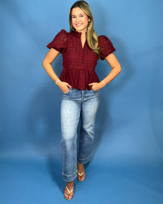 A woman modeling a burgundy eyelet lace short-sleeve blouse with puff sleeves and a ruffled hem, paired with light-wash blue jeans and beige sandal
