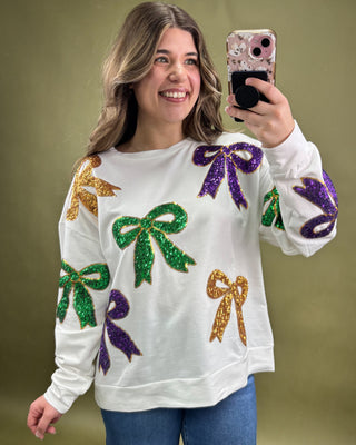 Woman wearing a white sweater with colorful bow designs, taking a selfie against a green background.