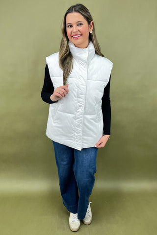 Woman wearing a white puffer vest over a black shirt and blue jeans on a beige background
