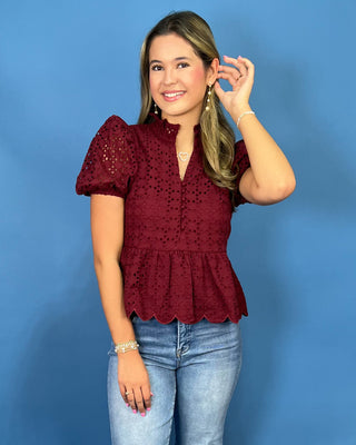 A woman modeling a burgundy eyelet lace short-sleeve blouse with puff sleeves and a ruffled hem, paired with light-wash blue jeans and beige sandal