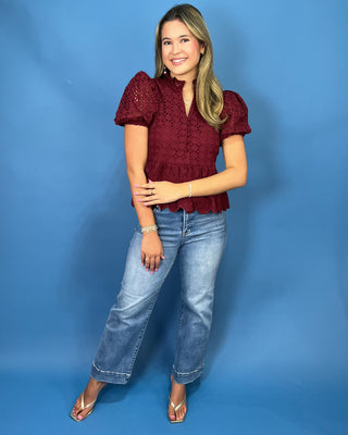 A woman modeling a burgundy eyelet puff-sleeve blouse with a ruffled hem, paired with light-wash blue jeans and metallic sandals