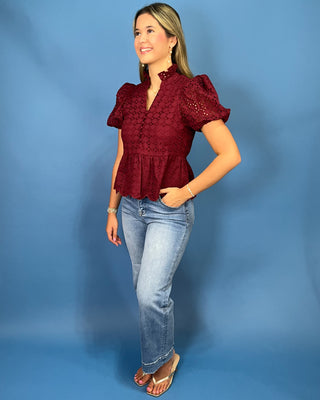 A woman modeling a burgundy eyelet lace peplum top with puff sleeves, paired with light-wash blue jeans and brown sandals
