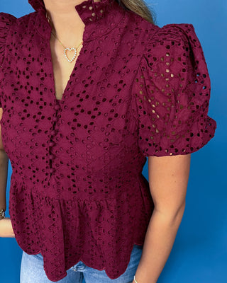 A woman wearing a burgundy eyelet lace blouse with puff sleeves and a ruffled hem, paired with blue jeans