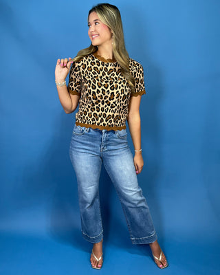 A woman modeling a short-sleeve leopard print top with brown trim, paired with light-wash blue jeans and beige sandals