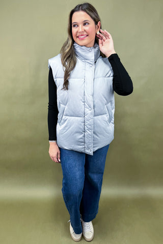 Woman wearing a light blue puffer vest over a black long-sleeve shirt and blue jeans on a beige background