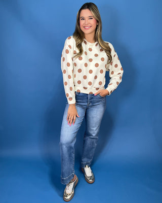 A woman modeling a cream long-sleeve top with brown polka dots, paired with blue jeans and metallic sneakers