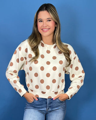A woman modeling a cream-colored long-sleeve sweatshirt with brown polka dots, paired with blue jeans